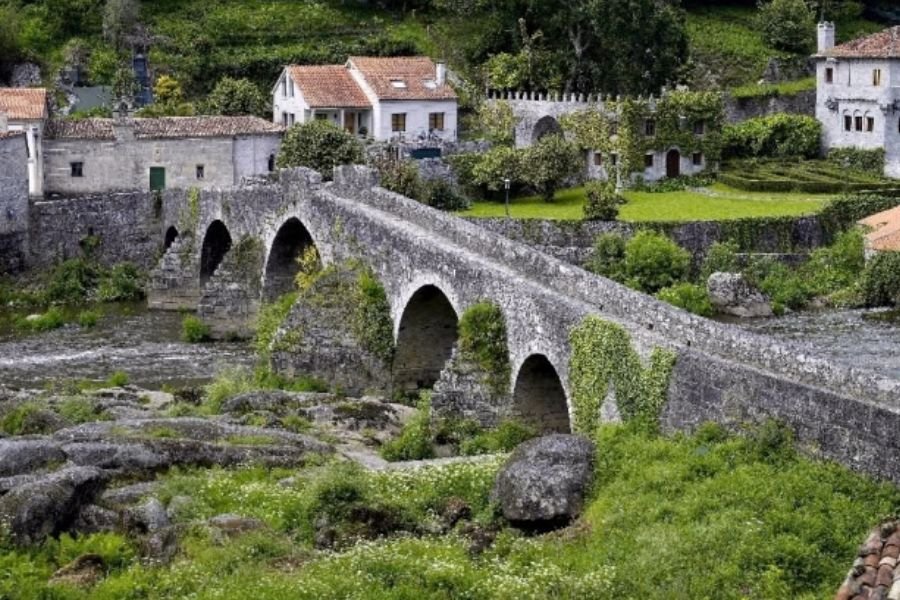 A ponte medieval de pedra de Ponte Maceira sobre o rio Tambre, com casas de pedra e um pazo ao fundo