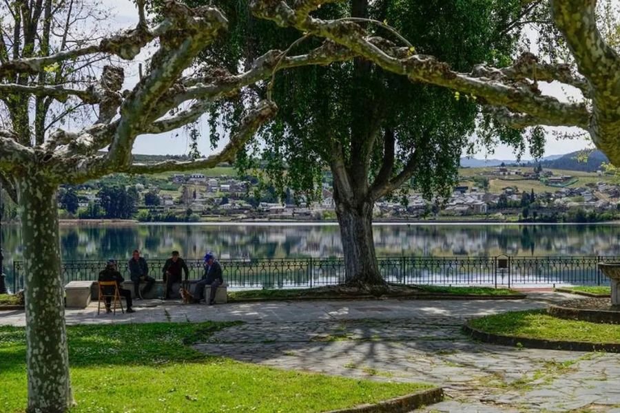 Eine schattige Uferpromenade in A Rúa de Valdeorras, mit Einheimischen auf einer Bank und dem Ort, der sich im ruhigen Wasser spiegelt