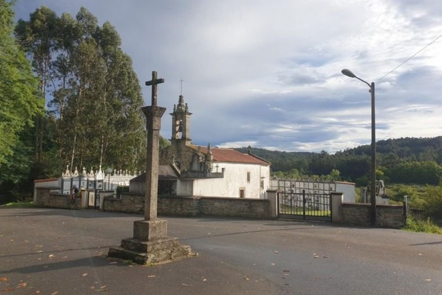 Ein hohes steinernes Wegkreuz vor einer weißen Dorfkirche und ihrem ummauerten Friedhof