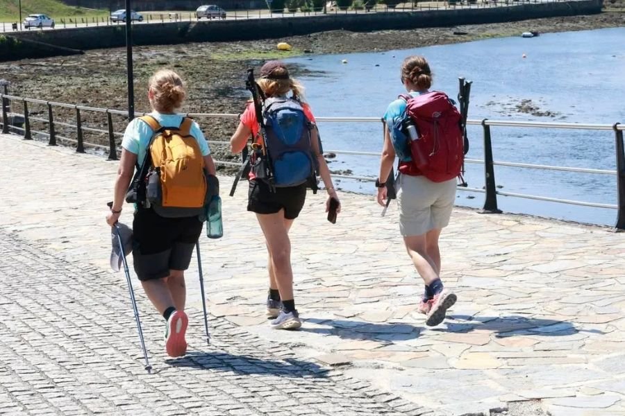 Drei Pilger mit Rucksäcken und Wanderstöcken auf einer steinernen Uferpromenade an einer Ría