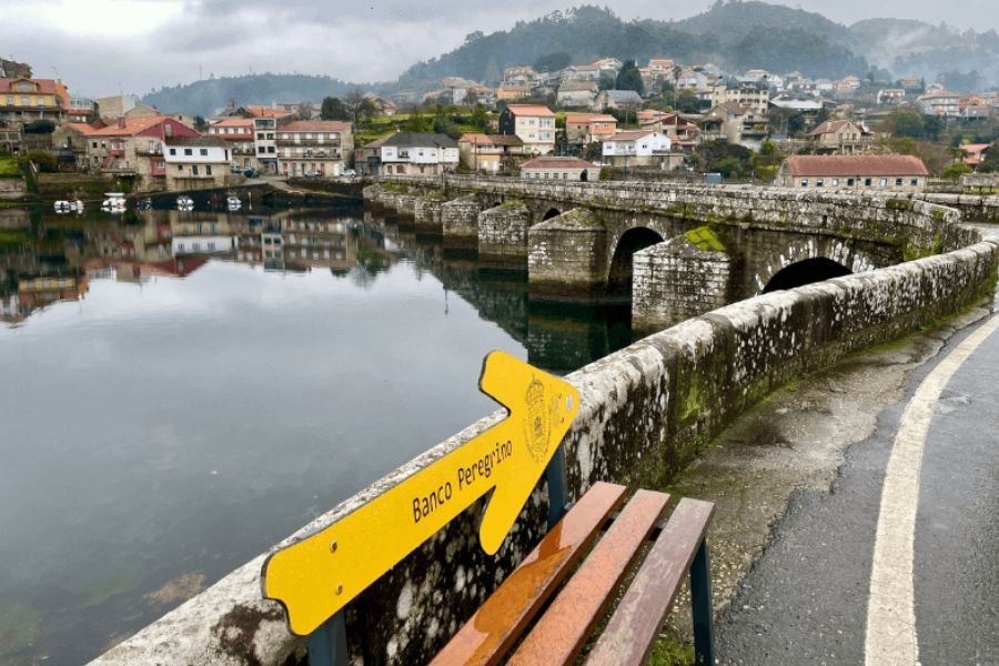 Ein gelber Pfeil mit der Aufschrift „Banco Peregrino“ auf einer Bank neben der mittelalterlichen Brücke Ponte Sampaio über den Verdugo