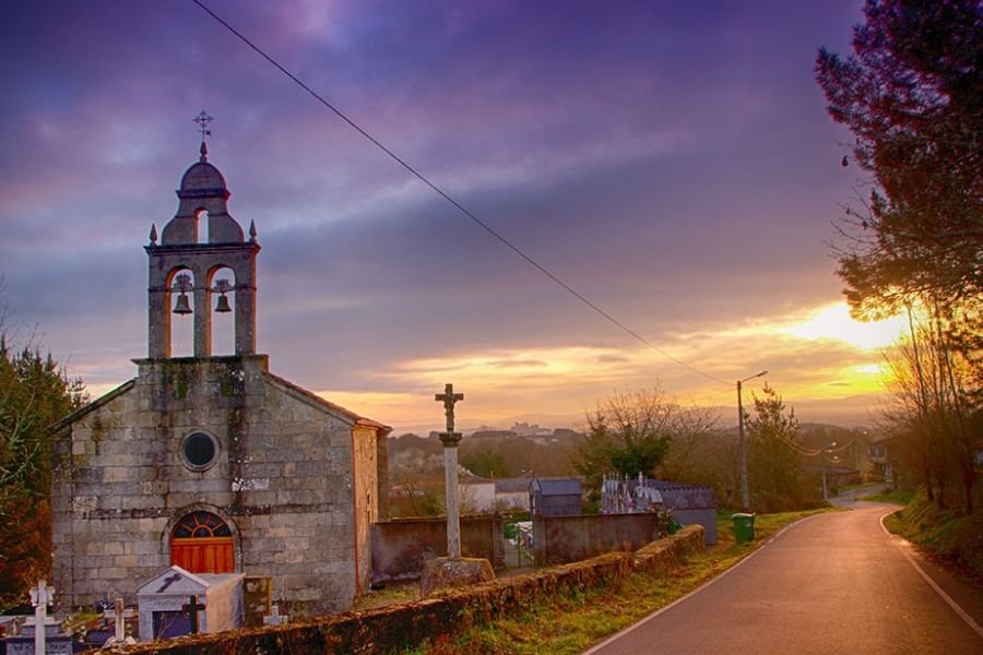 Eine kleine Steinkirche mit Glockengiebel, ein Steinkreuz und ein Dorffriedhof an einer Landstraße bei Sonnenuntergang, in der Gegend von Quiroga