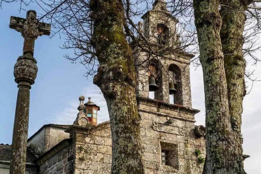 Il campanile in pietra della chiesa di Miraz e un’antica croce, tra alberi coperti di muschio