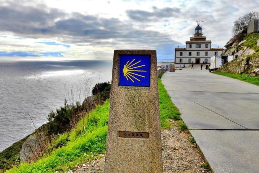 Il cippo del Cammino che indica Km 0,000 a Capo Fisterra, con il faro e l’oceano sullo sfondo