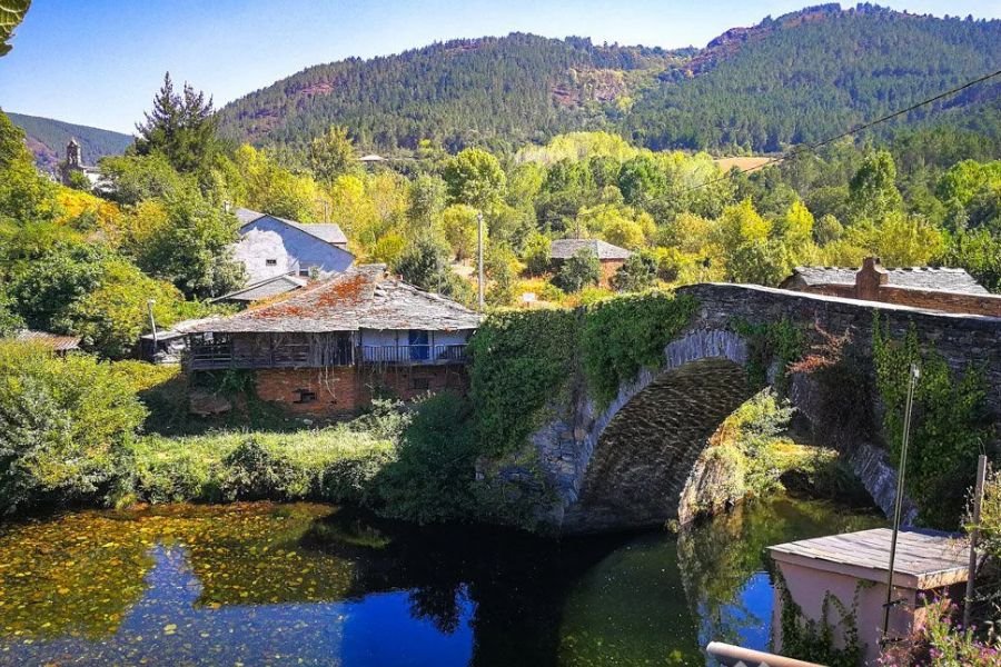 Un ponte ad arco in pietra coperto d’edera su un fiume calmo ad A Pobra do Brollón, con case dai tetti di ardesia e colline boscose sullo sfondo