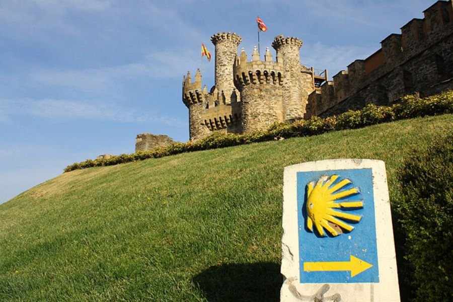 Las torres y almenas del Castillo de los Templarios de Ponferrada sobre una ladera cubierta de pasto, con un mojón del Camino con la concha de vieira en primer plano