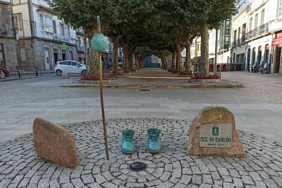 Un par de botas de bronce y un bordón de peregrino con una vieira en una plaza de piedra en Cee, junto a una piedra con la inscripción Cee no Camiño