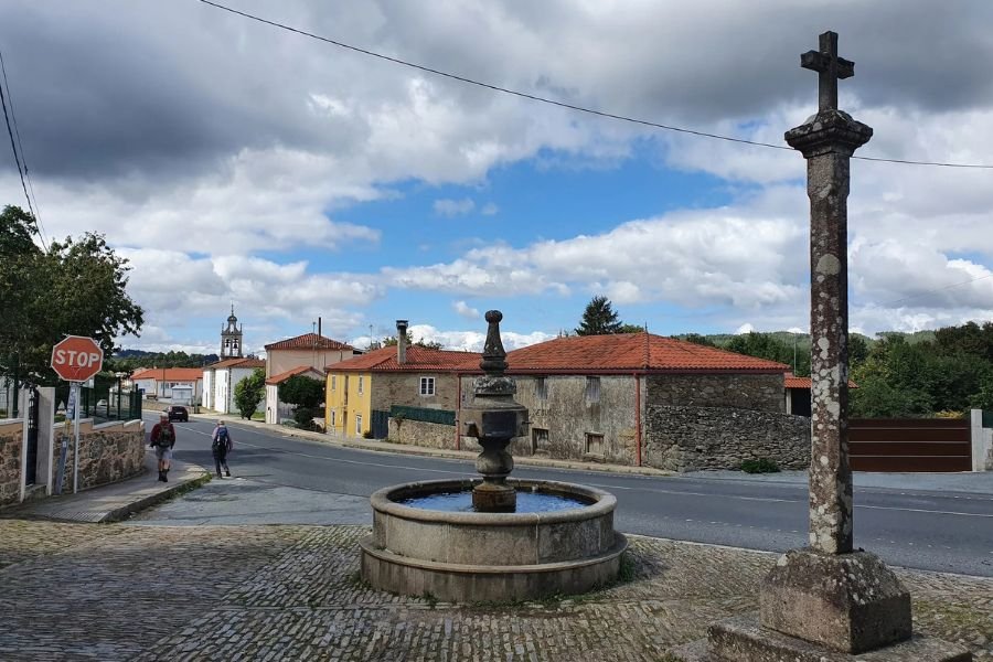 Un crucero de piedra y una fuente en la plaza de una aldea gallega, con peregrinos siguiendo su camino hacia Santiago