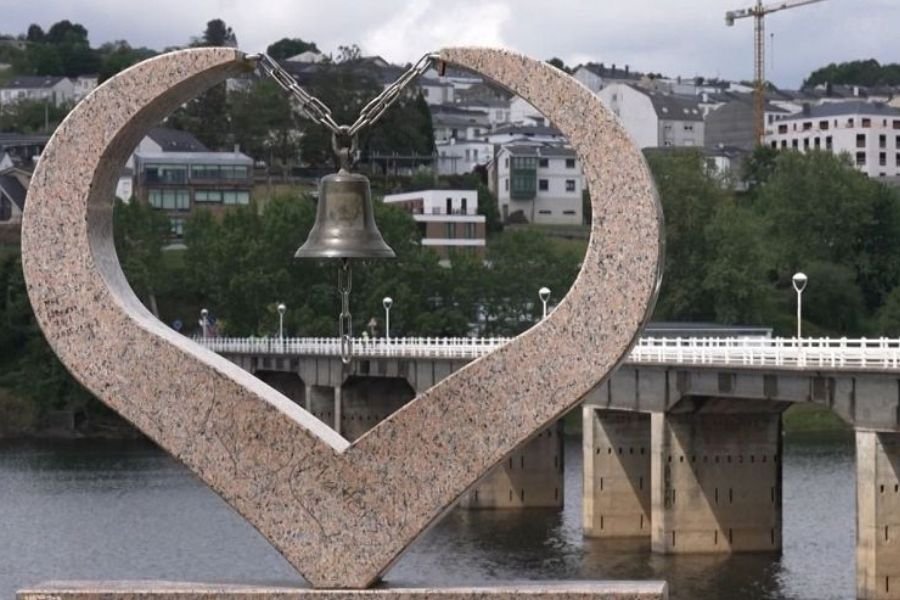 A escultura de granito em forma de coração com um sino em Portomarín, com a ponte e a vila ao fundo