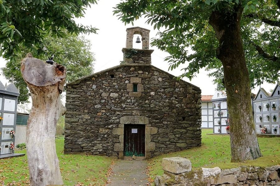 The small rustic stone church of San Lourenzo de Bruma, with a single bell gable, between trees and the village cemetery