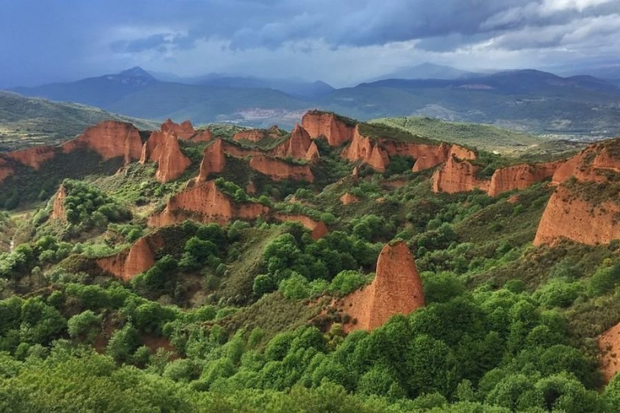 The red, jagged peaks of Las Médulas rising out of green chestnut woods, with mountains under storm clouds behind