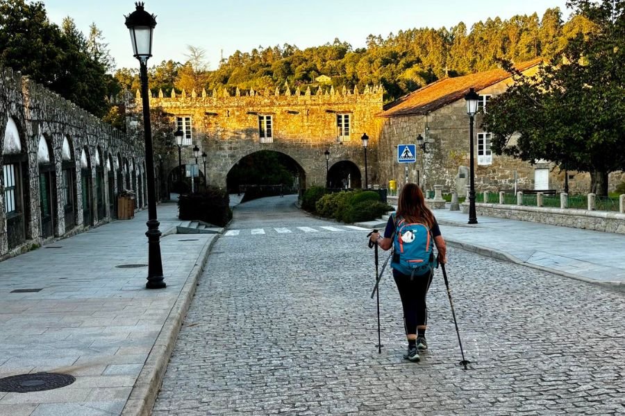 A pilgrim with trekking poles walking down a cobbled street toward a crenellated stone gateway in Negreira
